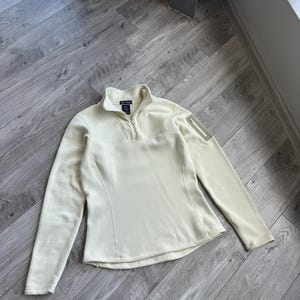 Könnte beinhalten: Cremefarbener Fleece-Pullover mit einem Viertel-Reißverschluss-Ausschnitt und langen Ärmeln. Er hat eine kleine Reißverschlusstasche am oberen linken Ärmel. Das Kleidungsstück liegt auf einem Holzboden.