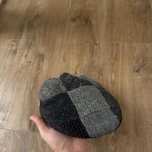 Może przedstawiać: Czapka typu newsboy z patchworku w odcieniach szarości, czerni i brązu. Czapka posiada różnorodne, teksturowane tkaniny, w tym wzory w jodełkę. Czapka trzymana jest w dłoni.
