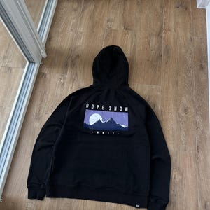 May include: Black hooded sweatshirt with a graphic on the back that reads "DOPE SNOW" above a mountain scene with a purple sky. The sweatshirt has a drawstring hood and ribbed cuffs and hem.