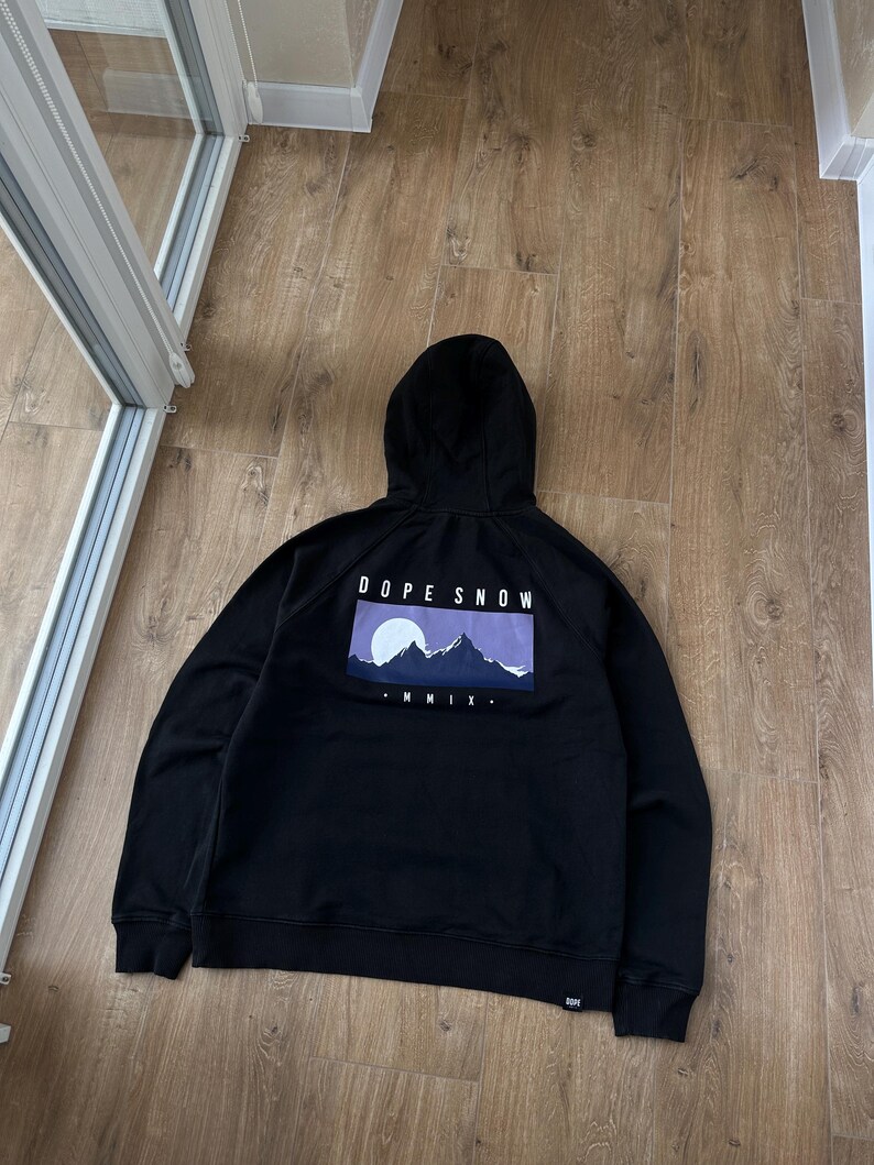 May include: Black hooded sweatshirt with a graphic on the back that reads "DOPE SNOW" above a mountain scene with a purple sky. The sweatshirt has a drawstring hood and ribbed cuffs and hem.
