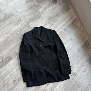 May include: A black pinstripe blazer with three buttons and two front pockets. The blazer is lying on a light gray wooden floor. The blazer has a notched lapel and buttoned cuffs. The garment is a classic style.