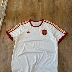 May include: A white short-sleeved t-shirt featuring a red Adidas logo on the right chest and the Spanish national football team crest on the left chest. The sleeves and shoulders are accented with red and yellow stripes along the top edges.