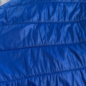 May include: Close-up of a bright blue quilted jacket. The jacket has a hood and horizontal stitching. The fabric has a slight sheen, and the overall appearance is smooth and sleek. The jacket is a vibrant shade of blue.