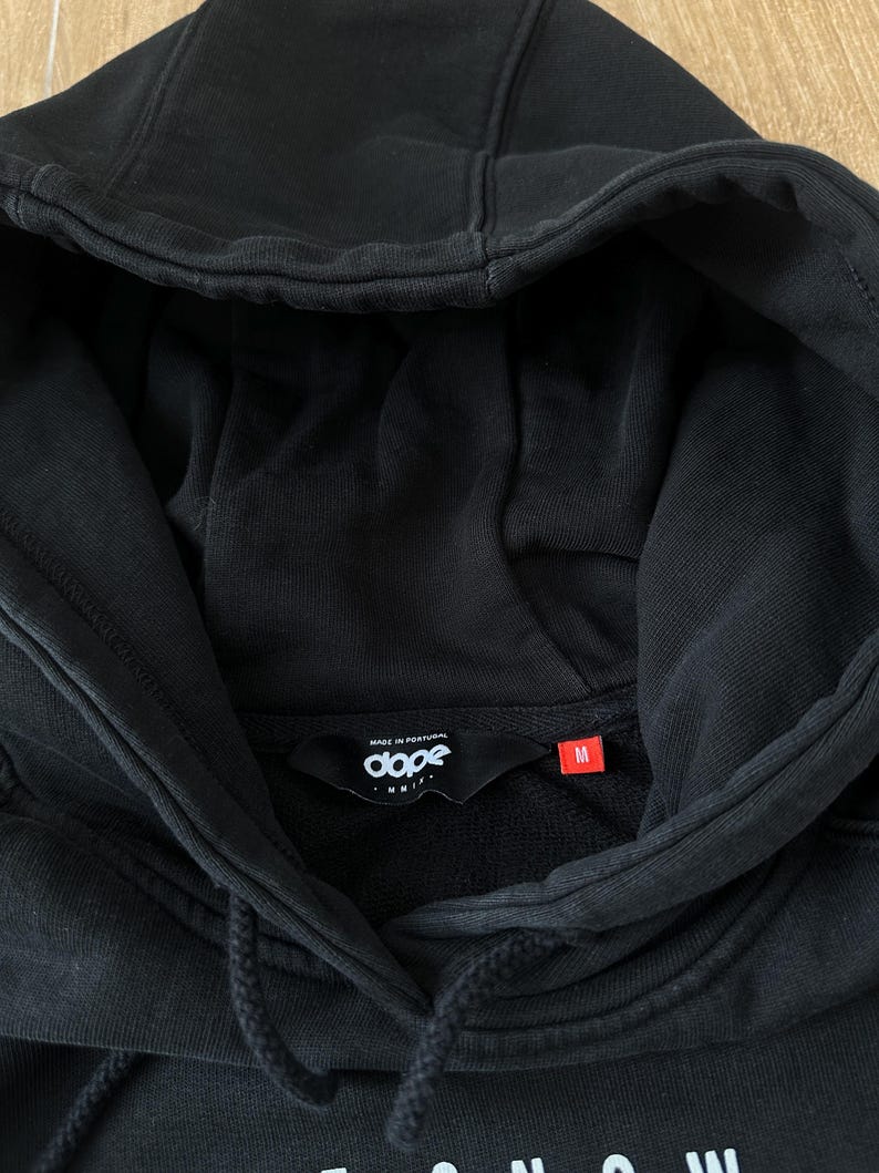 May include: Black hooded sweatshirt with a drawstring and a front pocket. The label inside the collar reads "dope" and "MADE IN PORTUGAL". The size tag is red and marked "M". The word "SNOW" is visible at the bottom of the sweatshirt.