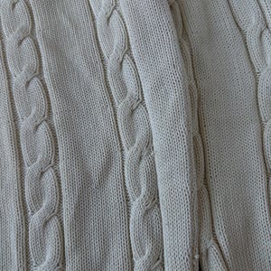 Puede incluir: Primer plano de un su&eacute;ter de punto trenzado color crema. El su&eacute;ter presenta un patr&oacute;n texturizado de puntadas de cable verticales y una textura de punto acanalado. La tela parece suave y c&oacute;moda.