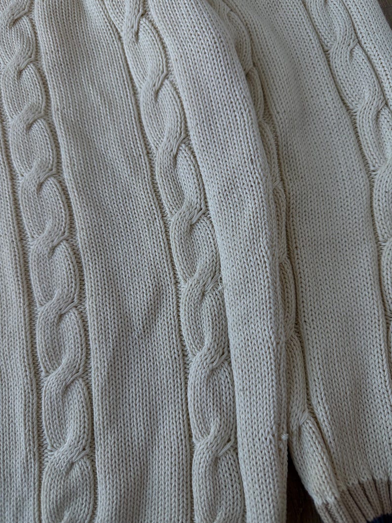 Puede incluir: Primer plano de un su&eacute;ter de punto trenzado color crema. El su&eacute;ter presenta un patr&oacute;n texturizado de puntadas de cable verticales y una textura de punto acanalado. La tela parece suave y c&oacute;moda.