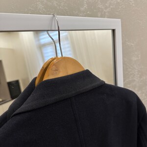 May include: A navy blue coat hanging on a wooden hanger. The coat is made of a dark, possibly wool, fabric. The collar is visible, and the coat appears to be well-tailored. The background includes a white framed mirror.