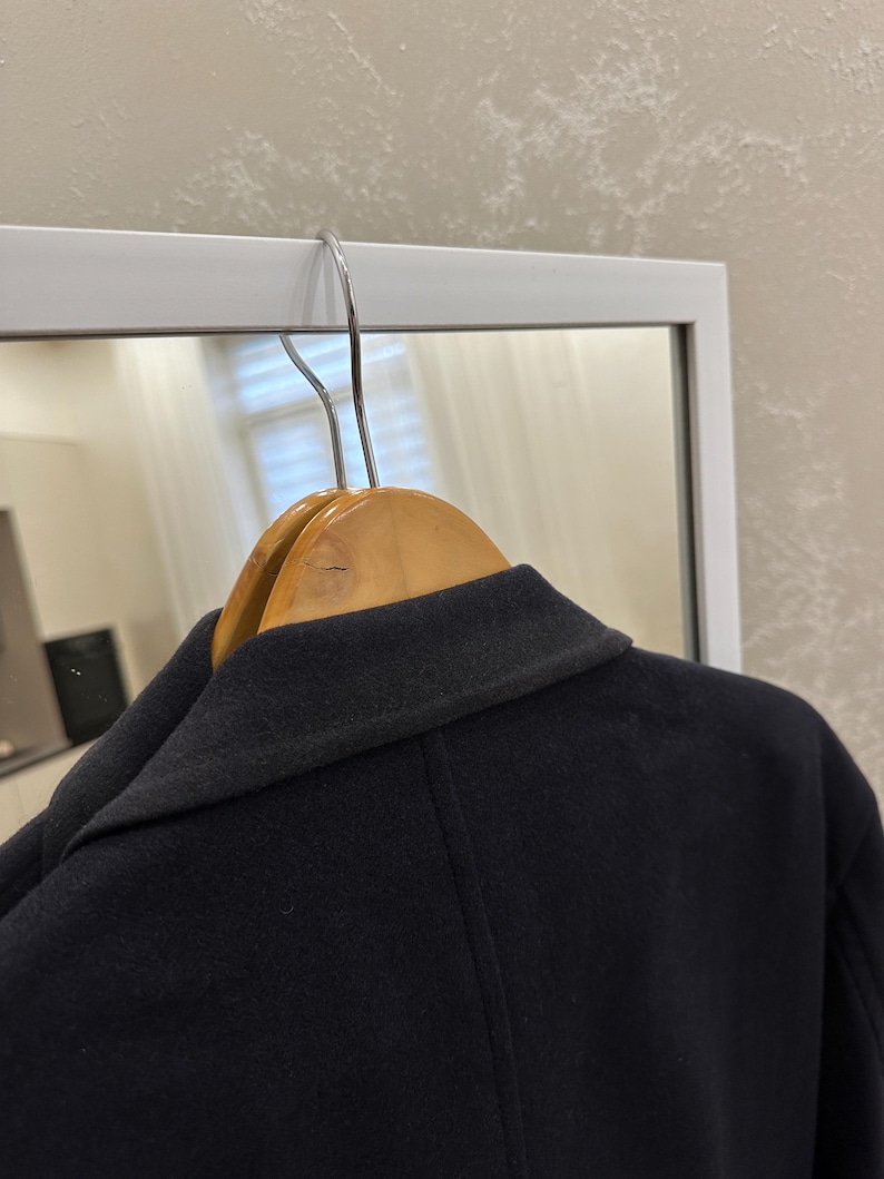 May include: A navy blue coat hanging on a wooden hanger. The coat is made of a dark, possibly wool, fabric. The collar is visible, and the coat appears to be well-tailored. The background includes a white framed mirror.