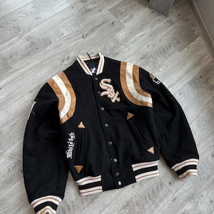 May include: Black varsity jacket with white and brown detailing. Features the text "White Sox" on the sleeve and the "SOX" logo on the chest. The jacket has a button-up front and striped cuffs and hem.