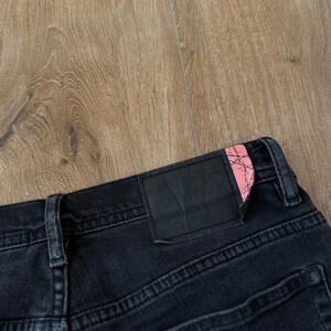 May include: Black denim jeans featuring a rectangular black leather-like patch on the back waistband. The patch has a distinct pink corner accent with a black cracked or marble-like pattern. The jeans include belt loops and visible stitching.