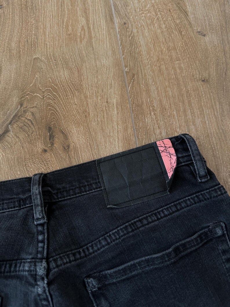 May include: Black denim jeans featuring a rectangular black leather-like patch on the back waistband. The patch has a distinct pink corner accent with a black cracked or marble-like pattern. The jeans include belt loops and visible stitching.