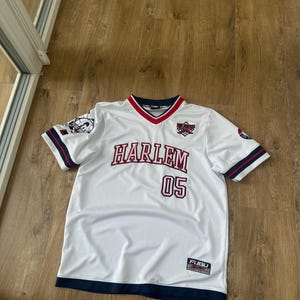 May include: A white V-neck jersey with navy blue and red trim on the collar and sleeves. The front features the text HARLEM and the number 05 in red and navy blue. Patches are on the sleeves, and a FUBU 05 92 label is at the bottom hem.