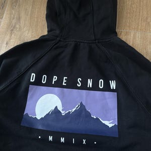 May include: Black hooded sweatshirt with the text "DOPE SNOW" above a graphic of a mountain range and a full moon. The graphic is in shades of purple and blue. The Roman numeral "MMIX" is below the graphic.