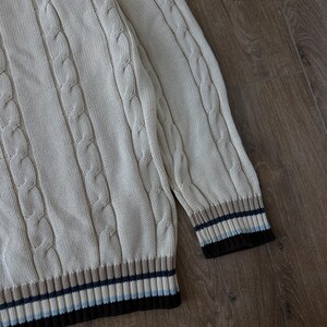 Puede incluir: Su&eacute;ter de punto trenzado color crema con dobladillo y pu&ntilde;os acanalados. Los pu&ntilde;os presentan rayas marrones, azul marino y celeste. El su&eacute;ter tiene un dise&ntilde;o cl&aacute;sico y texturizado.