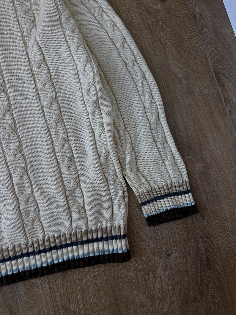 Puede incluir: Su&eacute;ter de punto trenzado color crema con dobladillo y pu&ntilde;os acanalados. Los pu&ntilde;os presentan rayas marrones, azul marino y celeste. El su&eacute;ter tiene un dise&ntilde;o cl&aacute;sico y texturizado.