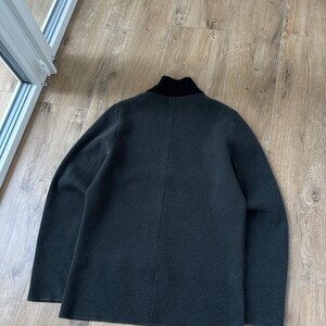 Op de afbeelding: Een donker antracietgrijze jas met lange mouwen en een effen zwarte opstaande kraag. Het kledingstuk is geconstrueerd met een zichtbare verticale naad die in het midden van de rug loopt en heeft een gladde, gelijkmatige textuur.