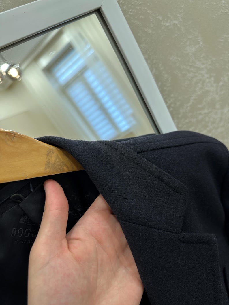 May include: A navy blue coat with a notched lapel is displayed on a wooden hanger. The coat's label reads "BOGO MILANO". The coat is made of a textured fabric.