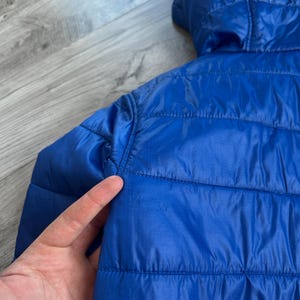 May include: A close-up of a bright blue quilted jacket with a hood. The jacket has horizontal stitching and a smooth, shiny texture. The hood is visible at the top, and the jacket appears to be lightweight.