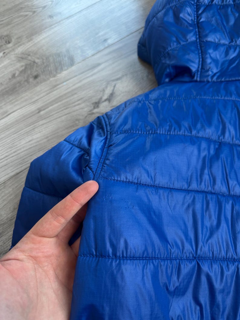 May include: A close-up of a bright blue quilted jacket with a hood. The jacket has horizontal stitching and a smooth, shiny texture. The hood is visible at the top, and the jacket appears to be lightweight.