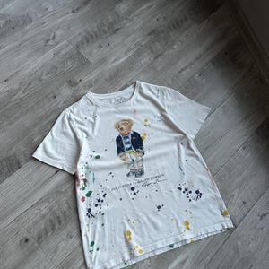 Op de afbeelding: Wit T-shirt met een Polo Bear-ontwerp met een blazer en een broek met patroon. Het shirt heeft kleurrijke verfspetters en de tekst "POLO BEAR by RALPH LAUREN".