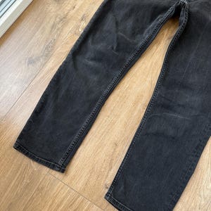 May include: A pair of black denim jeans featuring a faded wash, a button fly closure, and straight-cut legs. The fabric displays a worn-in texture with subtle whiskering patterns around the upper thigh area and reinforced stitching along the seams.