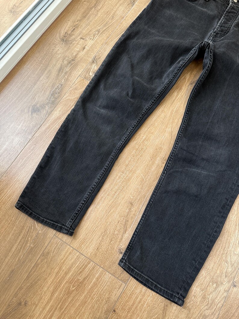 May include: A pair of black denim jeans featuring a faded wash, a button fly closure, and straight-cut legs. The fabric displays a worn-in texture with subtle whiskering patterns around the upper thigh area and reinforced stitching along the seams.