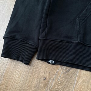 May include: Black hoodie with ribbed cuffs and hem. The word "DOPE" is printed on a small white label near the hem. The hoodie has a kangaroo pocket and a drawstring hood.