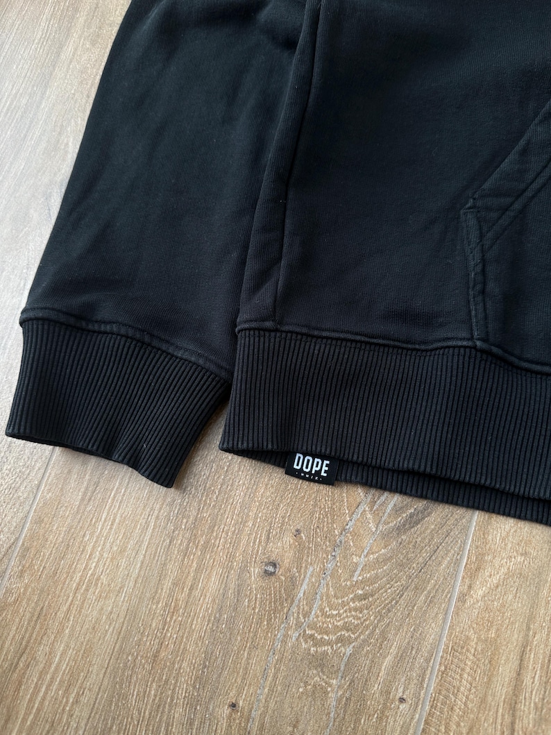 May include: Black hoodie with ribbed cuffs and hem. The word "DOPE" is printed on a small white label near the hem. The hoodie has a kangaroo pocket and a drawstring hood.