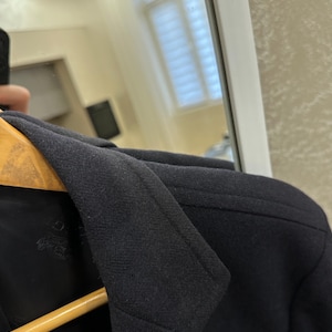 May include: Close-up of a black blazer with a notched lapel and a two-button closure. The blazer is hanging on a wooden hanger. The fabric appears to be a textured wool or wool blend.