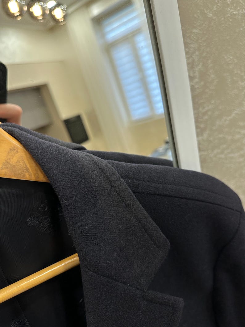 May include: Close-up of a black blazer with a notched lapel and a two-button closure. The blazer is hanging on a wooden hanger. The fabric appears to be a textured wool or wool blend.