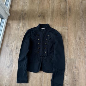 以下が含まれることがあります： ハイカラーとダブルブレストのフロントクロージャーを備えた黒いジャケット。真鍮色の丸いボタンが2列に計8個配置されています。フラップ付きのフロントポケットが2つあり、袖口にはステッチが施された長袖のデザインです。