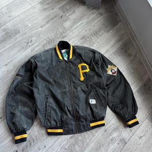 Könnte beinhalten: Schwarze Bomberjacke mit gelb-schwarz gestreiften Bündchen und Kragen. Die Jacke hat ein großes gelbes "P" auf der Brust und einen Pirates-Logo-Patch auf dem Ärmel. Die Jacke hat einen durchgehenden Reißverschluss und zwei Vordertaschen.