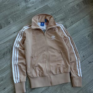 May include: A tan Adidas track jacket with white stripes down the sleeves and a white logo on the chest. The jacket has a full zipper, two front pockets, and a high collar. The jacket is made of a smooth, shiny material.