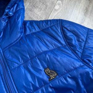 May include: Bright blue quilted puffer jacket with a hood. The jacket features a zipper closure and a small black and white owl logo patch on the chest. The fabric has a shiny, smooth texture.