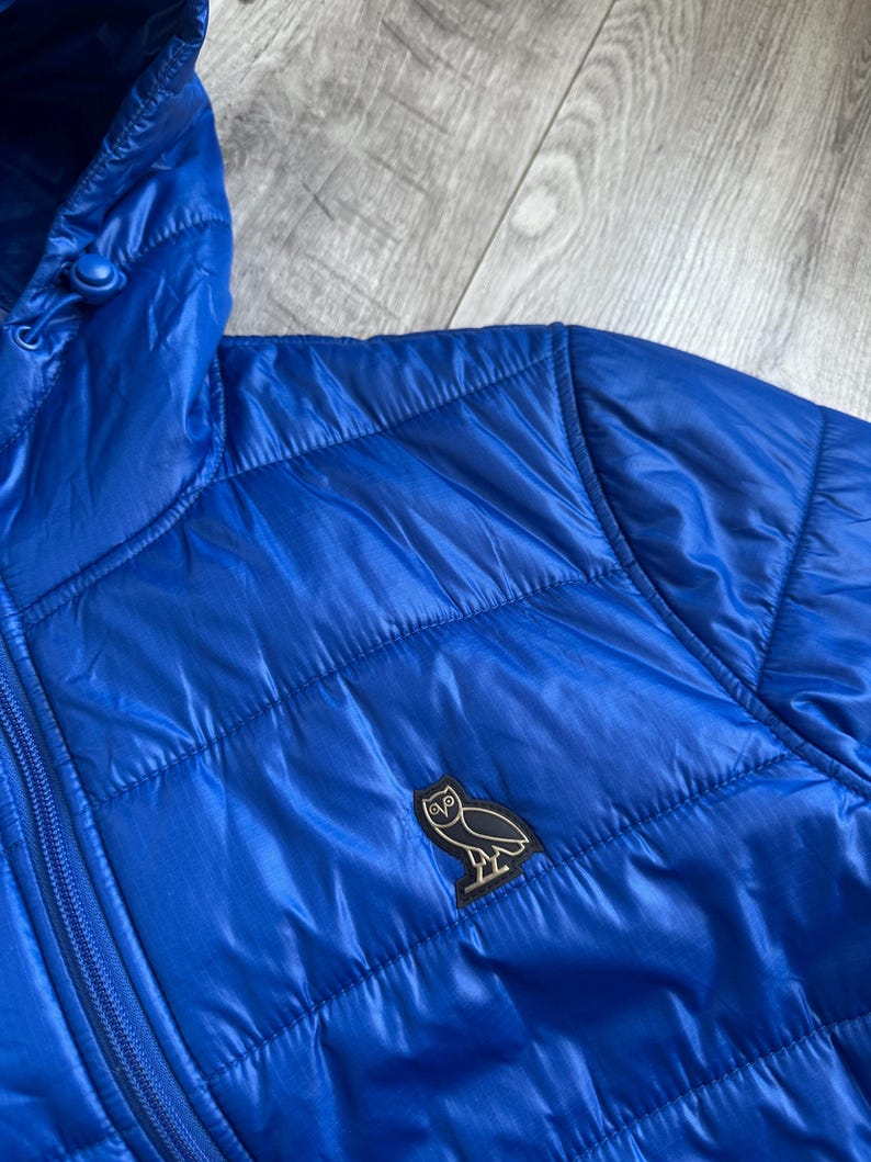 May include: Bright blue quilted puffer jacket with a hood. The jacket features a zipper closure and a small black and white owl logo patch on the chest. The fabric has a shiny, smooth texture.