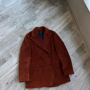 May include: A rust-colored corduroy blazer with a double-breasted design. The blazer features a notched lapel, two front pockets, and black buttons. The jacket is lying flat on a wooden floor.