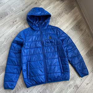 May include: A vibrant blue quilted jacket with a hood and a full-length zipper. The jacket features a small logo on the chest and a sleek, modern design. The jacket is made of a lightweight material.