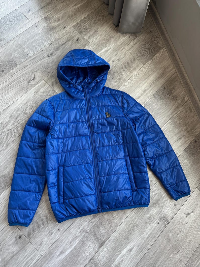 May include: A vibrant blue quilted jacket with a hood and a full-length zipper. The jacket features a small logo on the chest and a sleek, modern design. The jacket is made of a lightweight material.