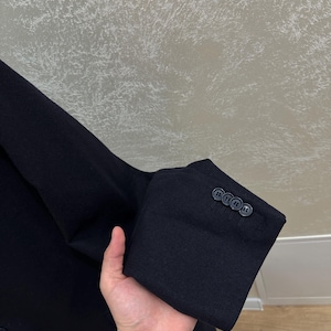 May include: Close-up of a black blazer sleeve with three dark buttons. The fabric appears to be a dark wool or wool blend. The sleeve is held up, showing the cuff and buttons.