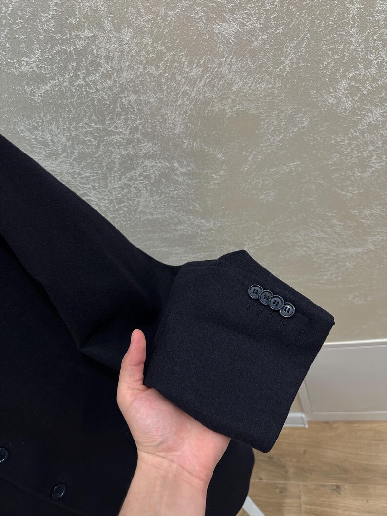 May include: Close-up of a black blazer sleeve with three dark buttons. The fabric appears to be a dark wool or wool blend. The sleeve is held up, showing the cuff and buttons.