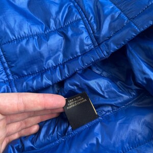 May include: Bright blue quilted jacket with a hood. The jacket has a shiny, smooth texture and visible stitching. A black tag with white text reads "CA# 57830 MADE IN CHINA".