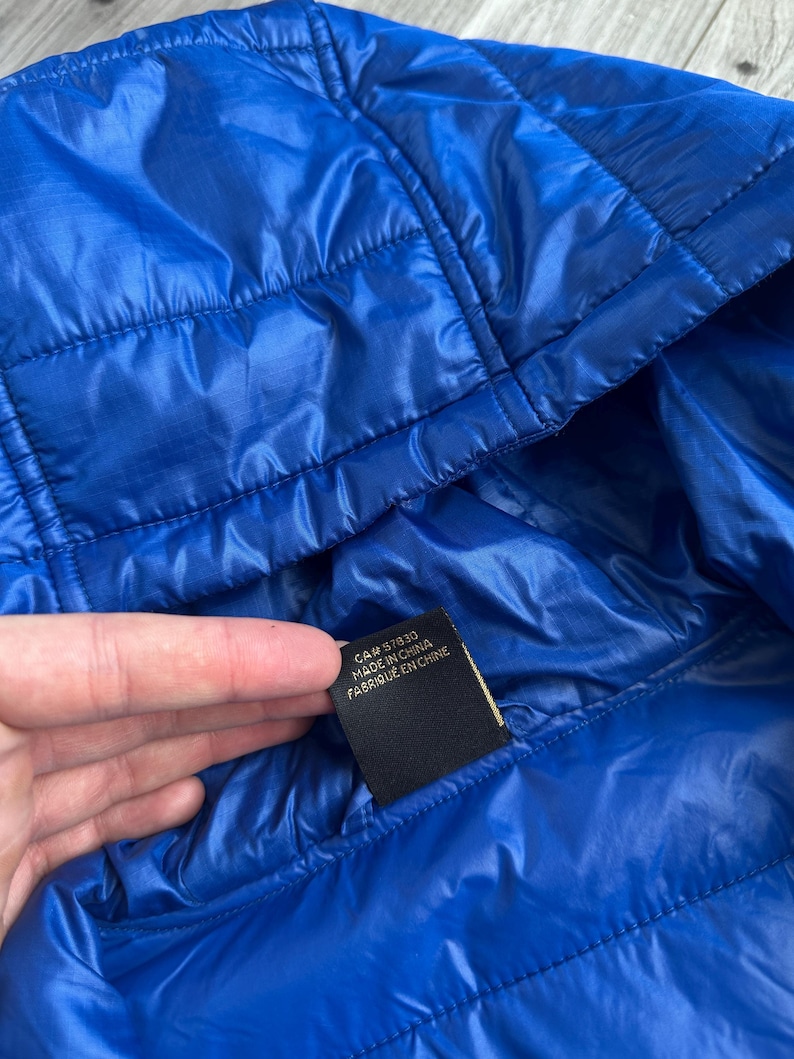 May include: Bright blue quilted jacket with a hood. The jacket has a shiny, smooth texture and visible stitching. A black tag with white text reads "CA# 57830 MADE IN CHINA".