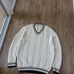 Puede incluir: Su&eacute;ter de punto trenzado color crema con cuello en V y ribetes a rayas. El su&eacute;ter presenta un dise&ntilde;o cl&aacute;sico de punto trenzado y pu&ntilde;os acanalados. Los ribetes incluyen rayas negras, marrones y azul marino. Una prenda casual y c&oacute;moda.