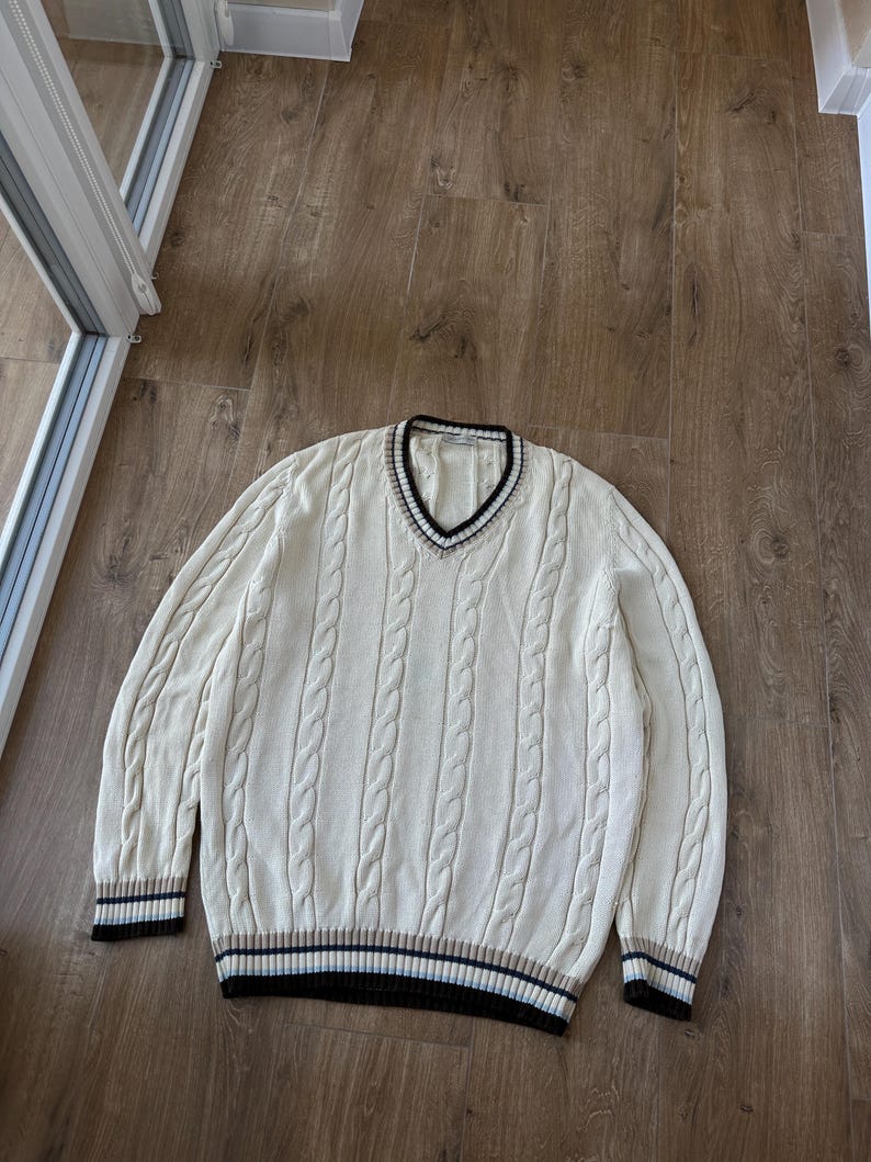 Puede incluir: Su&eacute;ter de punto trenzado color crema con cuello en V y ribetes a rayas. El su&eacute;ter presenta un dise&ntilde;o cl&aacute;sico de punto trenzado y pu&ntilde;os acanalados. Los ribetes incluyen rayas negras, marrones y azul marino. Una prenda casual y c&oacute;moda.