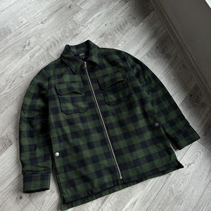 Puede incluir: Una camisa de franela a cuadros verde y negro con cierre de cremallera. La camisa tiene un bolsillo de parche en el pecho izquierdo y un bolsillo con solapa abotonada en el pecho derecho.