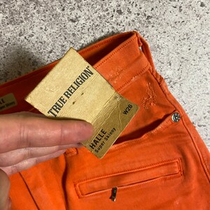 May include: A pair of orange jeans with a brown tag that says "True Religion" and a smaller tag that says "Halle Super Skinny" with the size W26.