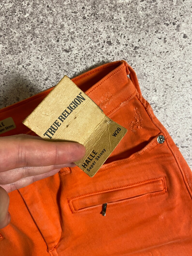 May include: A pair of orange jeans with a brown tag that says "True Religion" and a smaller tag that says "Halle Super Skinny" with the size W26.