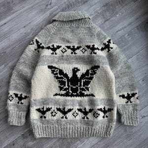 May include: A gray and cream-colored knit sweater featuring a large black eagle design on the back. The sweater has a shawl collar and is adorned with black bat motifs. The garment is made of a thick, textured yarn.