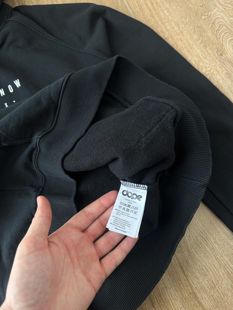 May include: Black hooded sweatshirt with the word "NOW" printed in white on the front. The sweatshirt has a ribbed hem and cuffs. A tag with the brand "dope" is visible.