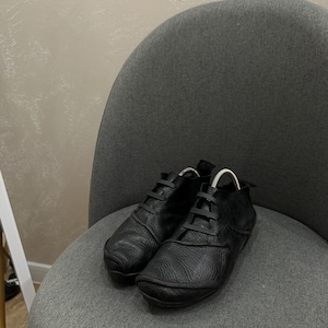 May include: A pair of black leather shoes with a textured surface, featuring a rounded toe, a low-cut ankle design, and a unique lacing system with wide, flat black laces that thread through integrated loops across the top of the footwear.